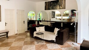 A spacious living room with a brown sofa, a small round table, and a ceiling fan. There are three arched windows and a painting on the wall. - Tropical Oasis 2 BR Villa Near Beach (Seminyak)