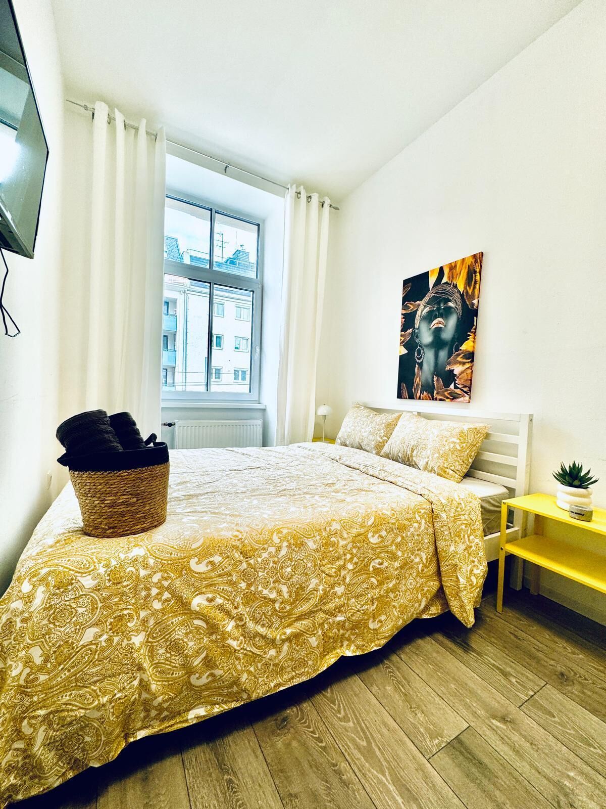 A bedroom with a bed, a window, a painting, a bedside table, and a basket.