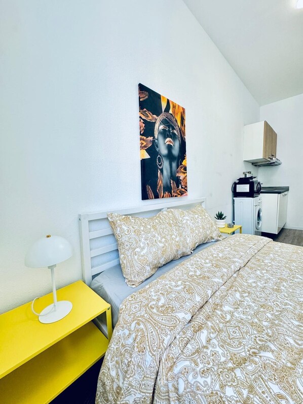 A bedroom with a bed, a nightstand, a lamp, a painting, a washing machine, and a cabinet. - Cozy and Affordable Palace Studio (Vienna)