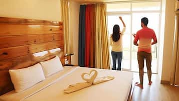 A hotel room with a wooden headboard, a bed with a heart-shaped towel, and a view of the outside through a sliding door.