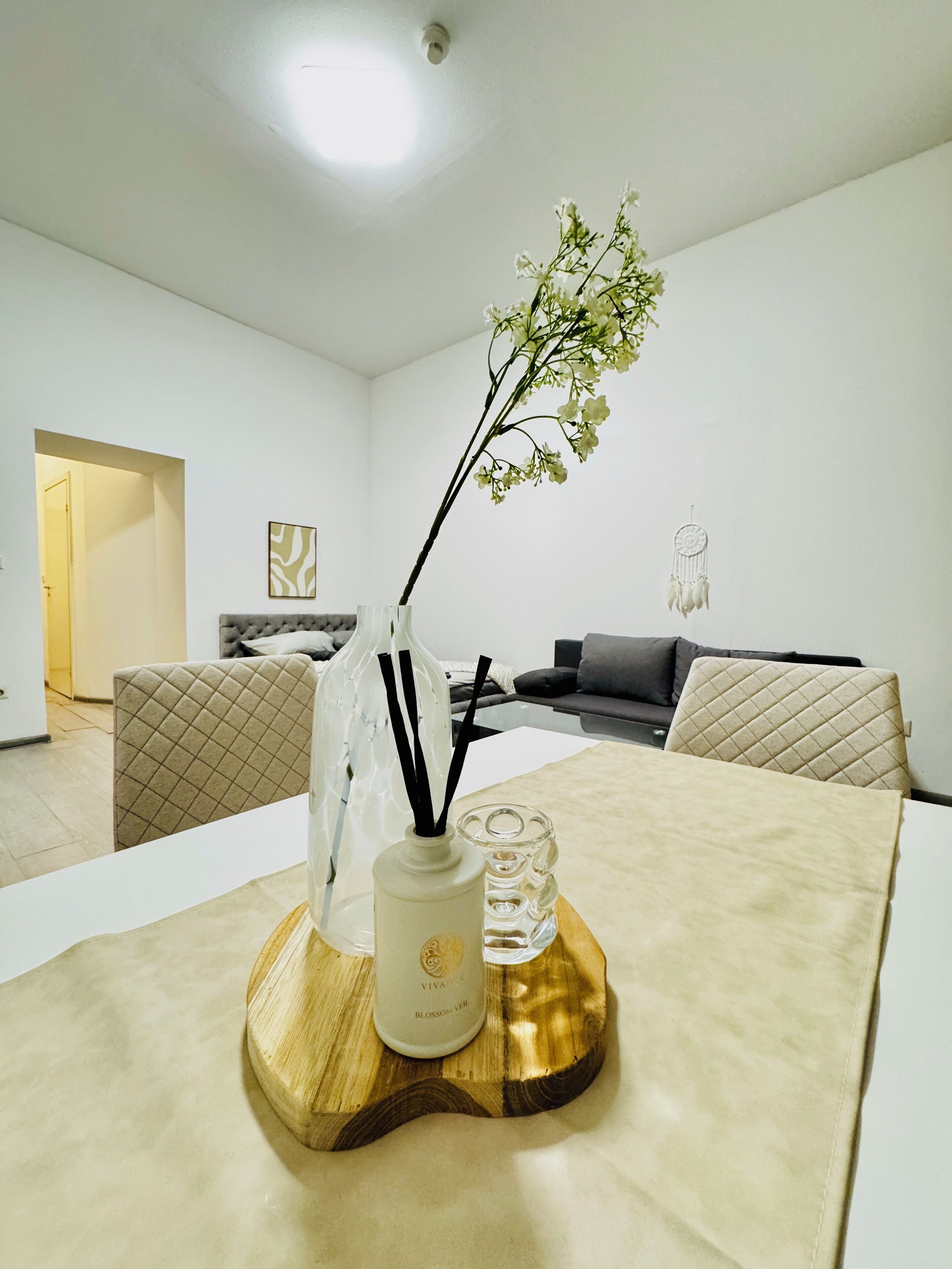 Una sala moderna con una mesa de comedor dispuesta, adornada con un florero, velas y una botella. Un sofá y un sillón se observan al fondo.