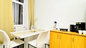 Una habitación pequeña y luminosa con una mesa blanca, dos sillas, un armario amarillo y una ventana con cortina.