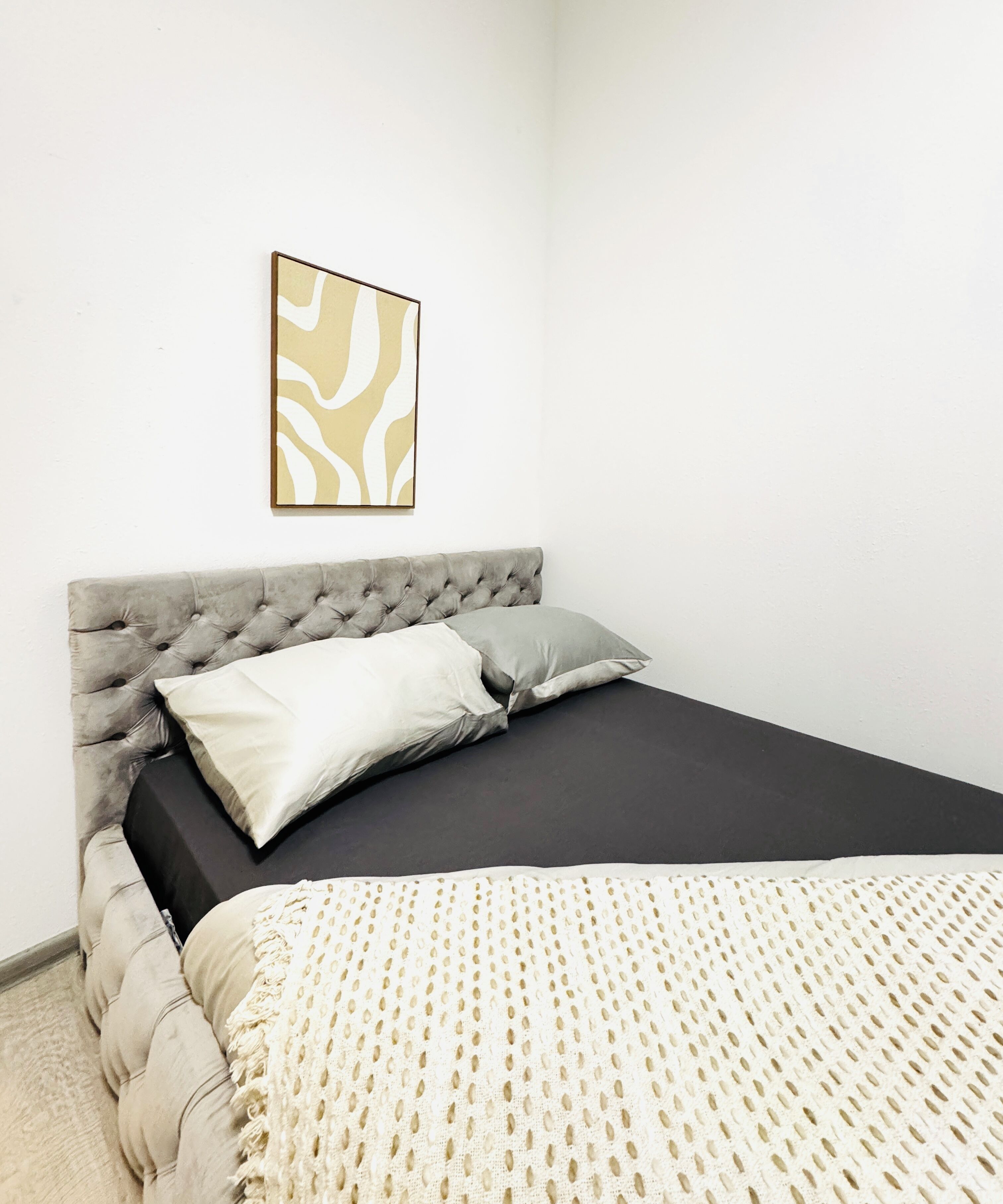 Un dormitorio con cabecera capitonada, colchón oscuro y manta blanca y beige con estampado.