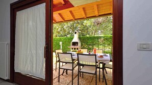 A patio area with a table and chairs, a grill, and a view of a garden. - Holiday home with air conditioning and terrace (Lignano Sabbiadoro)