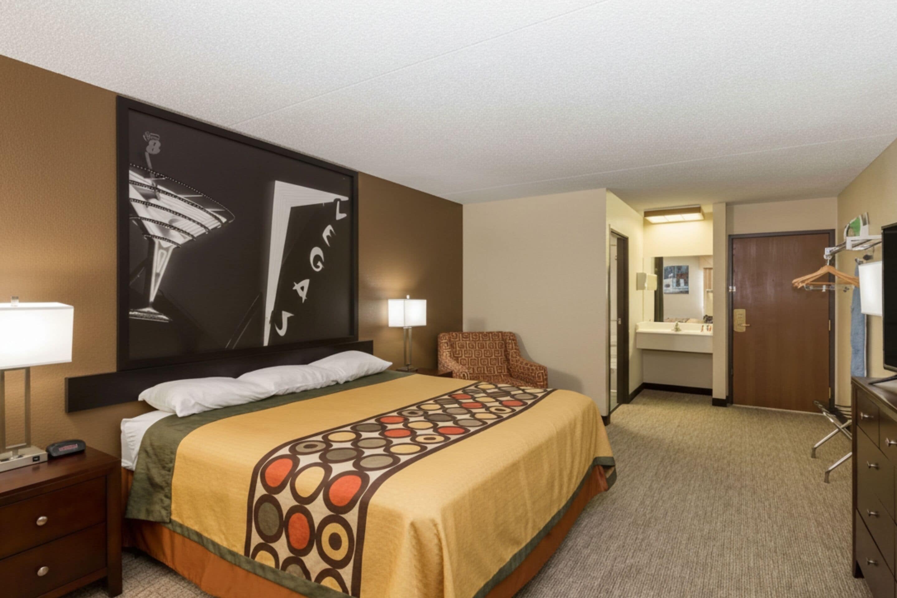 A hotel room with a bed, bedside table, lamp, and a large framed picture on the wall.