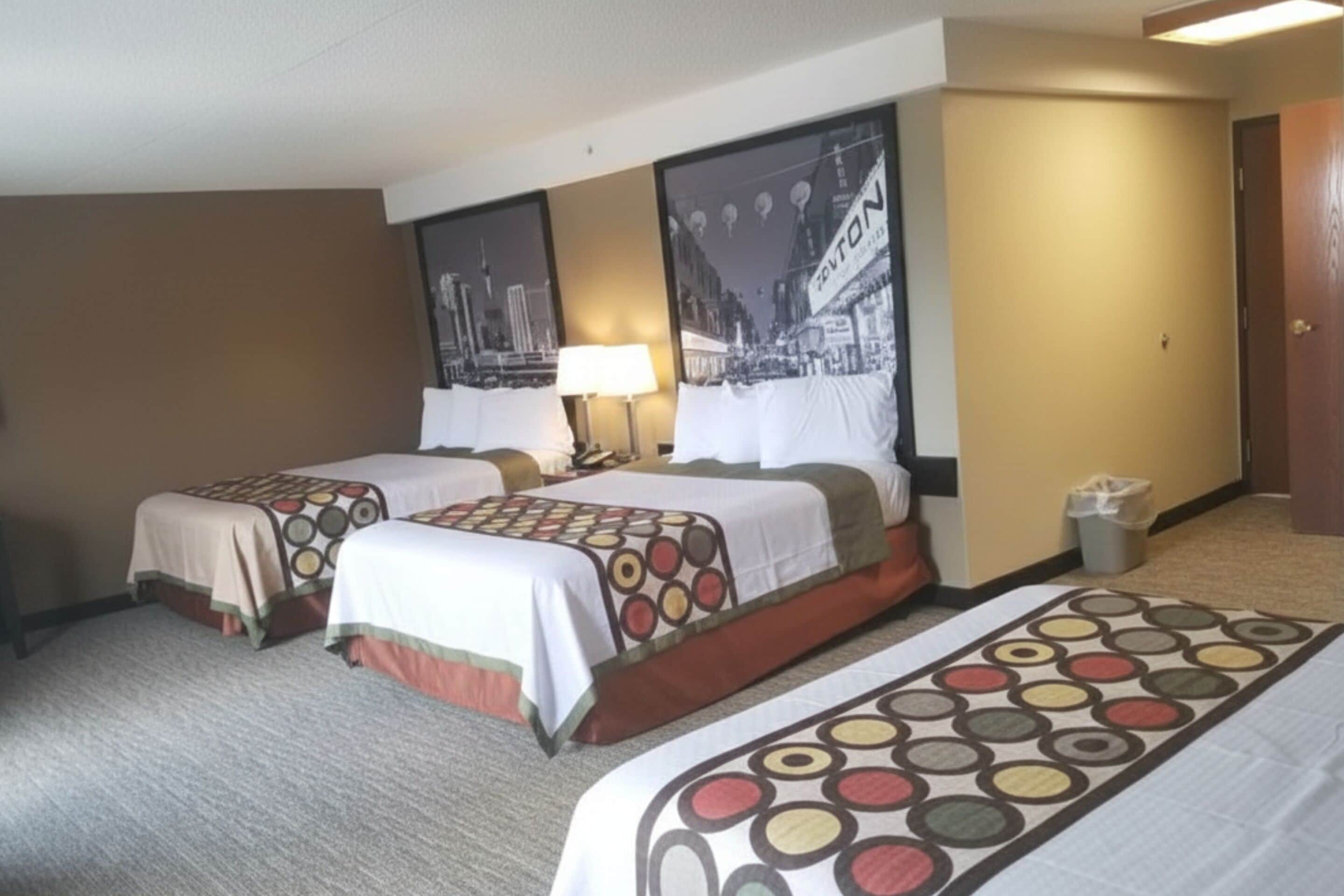 Habitación de hotel con dos camas, cada una con una colcha estampada, una mesita de noche con lámpara y un cuadro enmarcado con una vista urbana.