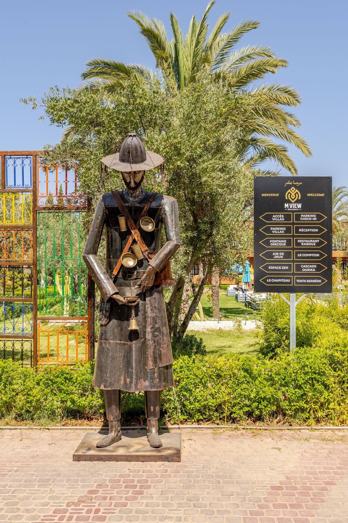 Une statue représentant une figure coiffée d’un chapeau et tenant un bâton, placée devant un panneau indiquant diverses commodités.