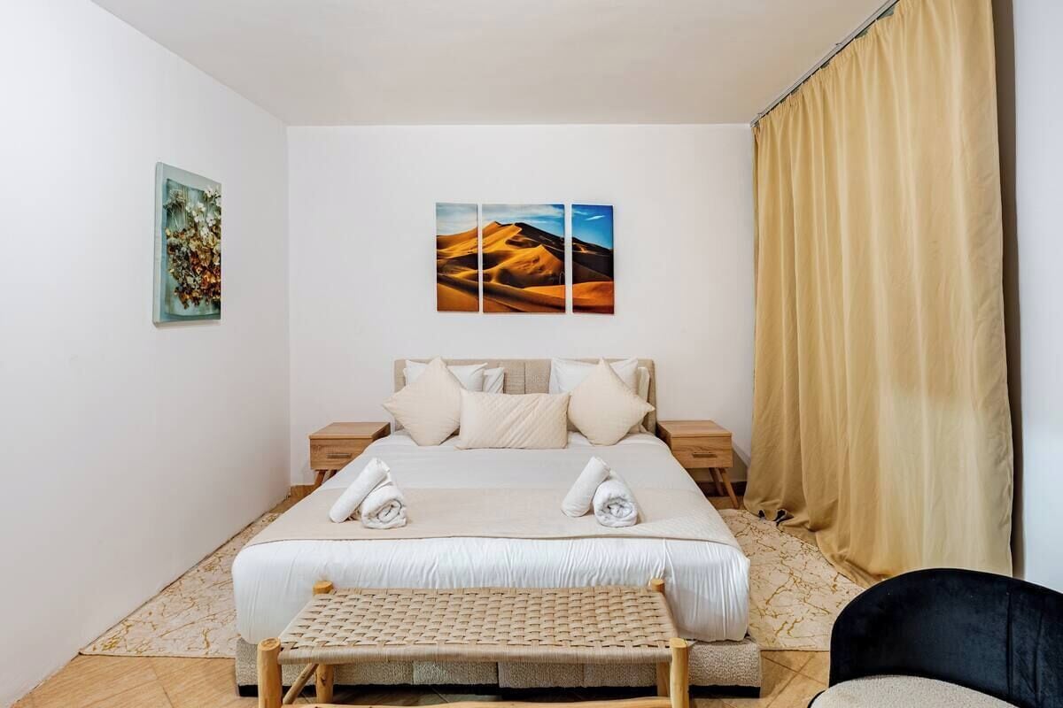 A hotel room with a bed, a bench, a chair, a nightstand, and artwork on the wall.