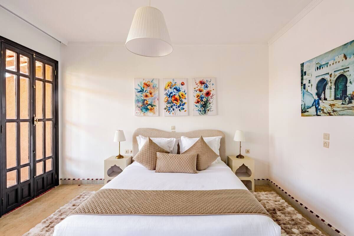 A bedroom with a bed, two bedside tables, a hanging lamp, and three floral paintings on the wall.