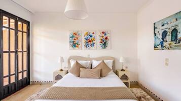 A bedroom with a bed, two bedside tables, a hanging lamp, and three floral paintings on the wall.