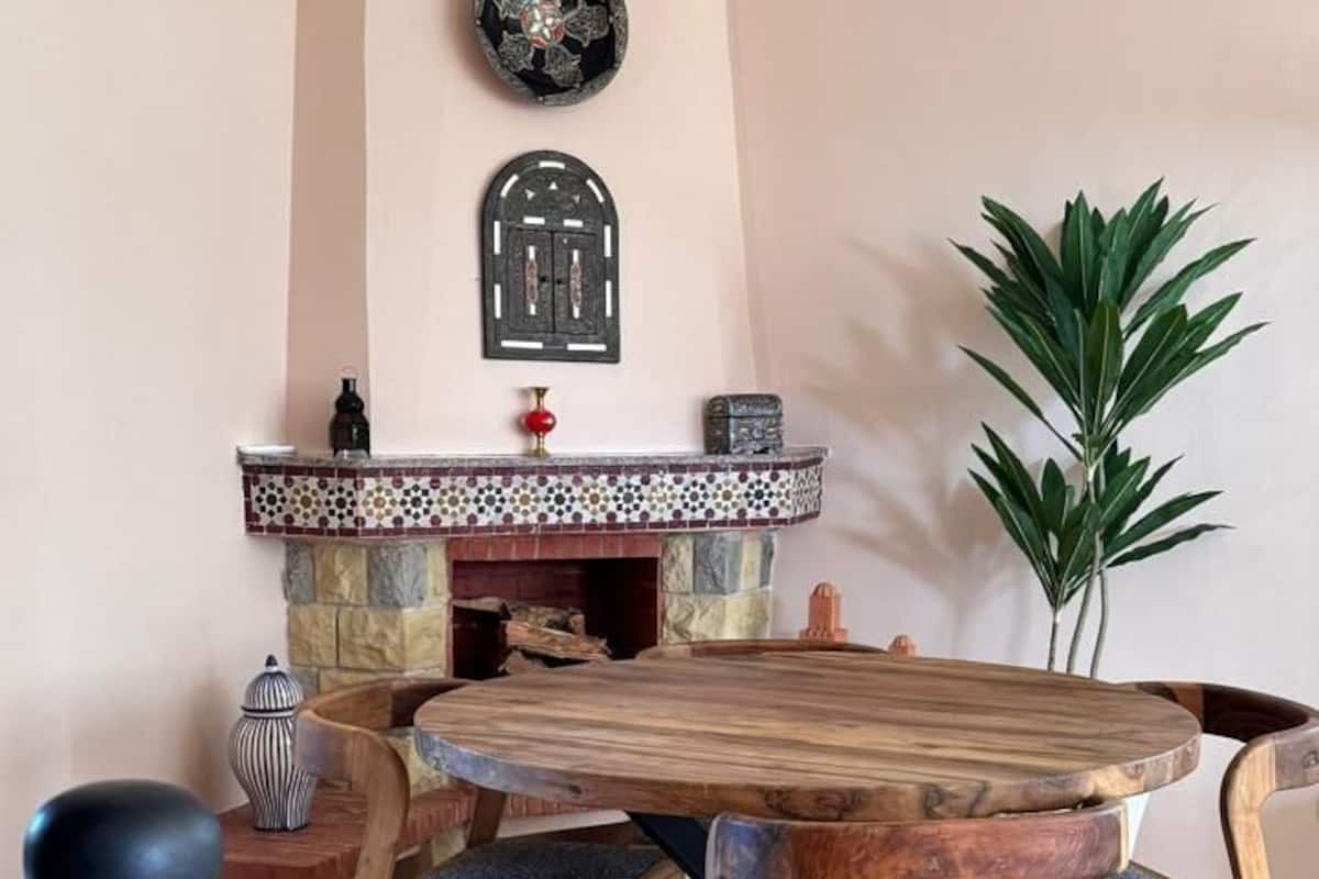 A dining area with a round wooden table, six chairs, a mosaic fireplace, and a potted plant.