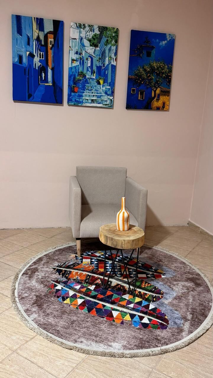 Ein Zimmer mit einem grauen Sessel, einem runden Holztisch mit einer Vase und einem farbenfrohen, geometrischen Teppich.