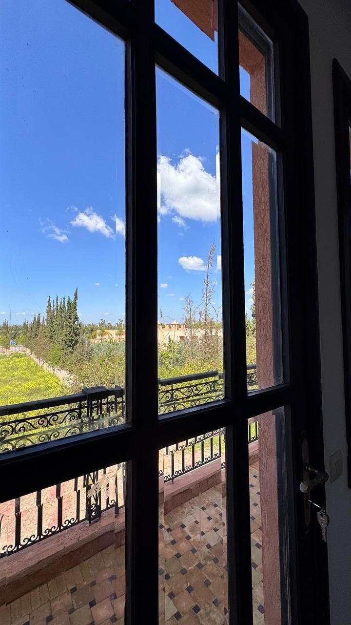 A view through a window with a balcony, featuring a clear sky, greenery, and distant buildings.