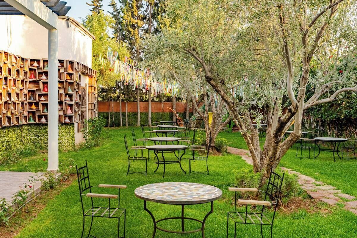 Un espacio al aire libre con mesas y sillas, rodeado de vegetación y un edificio con fachada singular.