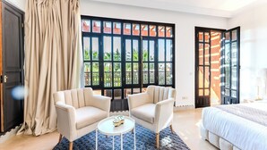 A bedroom with a bed, two armchairs, a small round table, and a large window with a view of greenery. - MVIEW Villas (Marrakech)