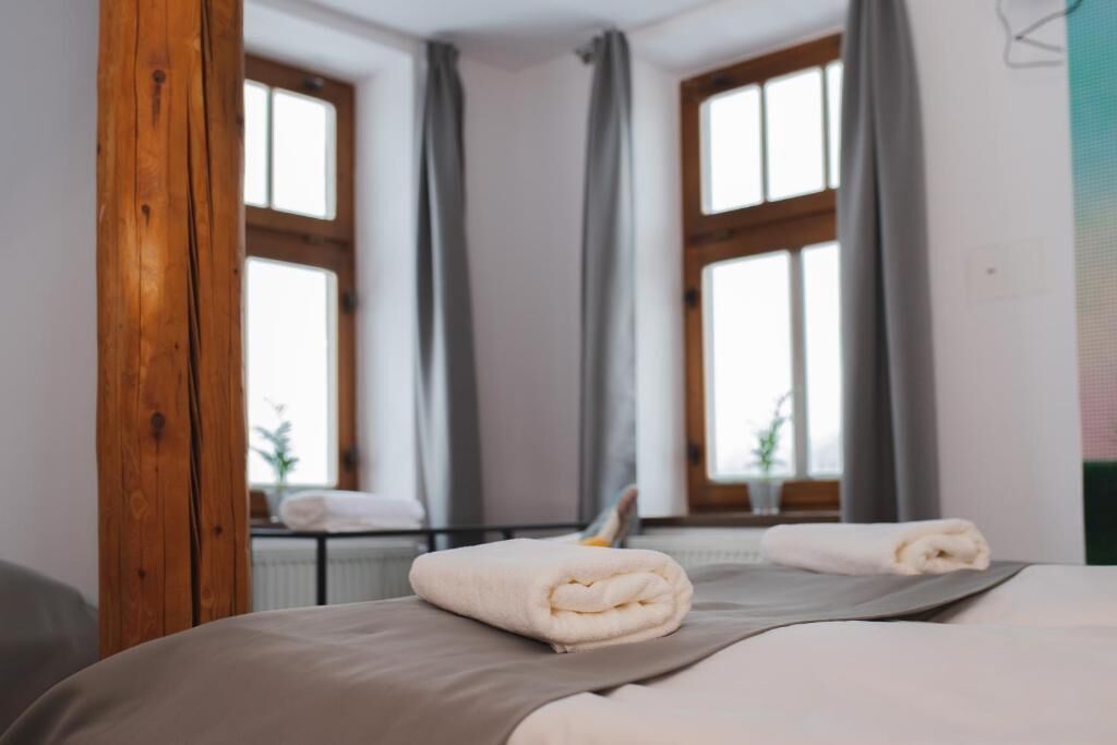 A hotel room with two beds, grey bedding, wooden beams, large windows with grey curtains, and a view of the outdoors.