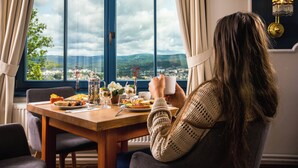 Ruokapöytä, josta avautuu näkymä maisemaan ikkunan kautta, pöydän ääressä istuva henkilö ja seinällä oleva kattokruunu.