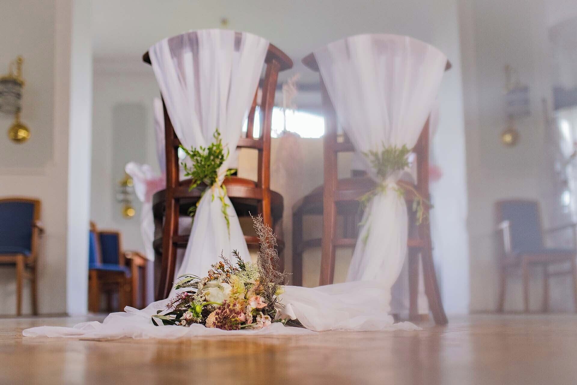 Ein Zimmer mit Holzboden, einem Stuhl mit weißem Stoffüberwurf und einem Blumenstrauß auf dem Boden.
