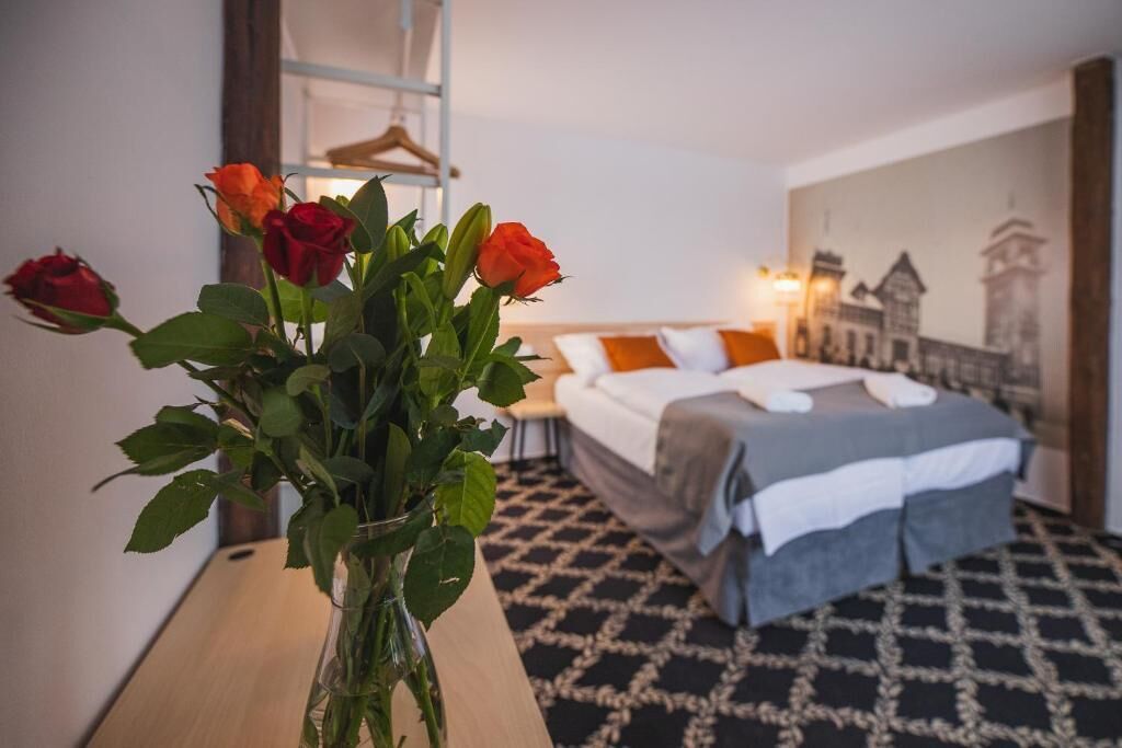 A hotel room with a bed, a nightstand, a vase with roses, and a wall with a cityscape painting.