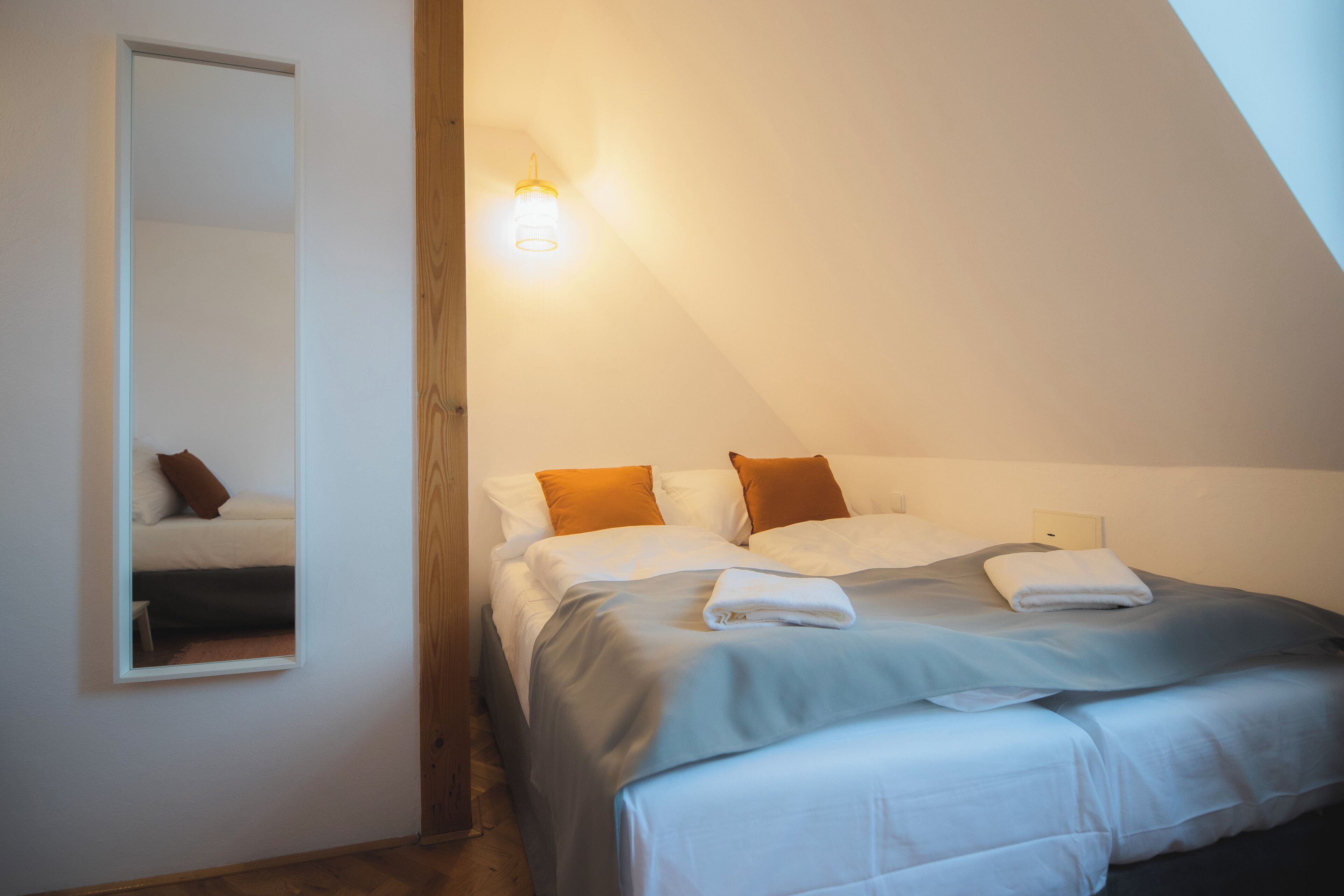 Quarto com teto inclinado, cama com roupa de cama branca e azul, duas almofadas, mesa de cabeceira e luminária fixa na parede.