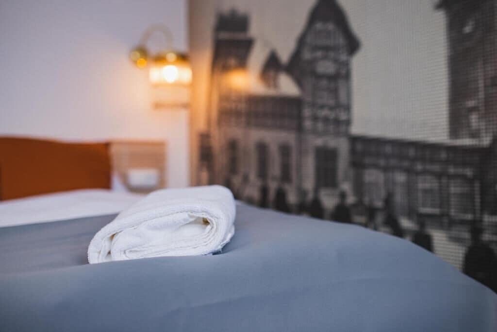 A neatly made bed with a folded white towel, a headboard, and a wall with a building mural.