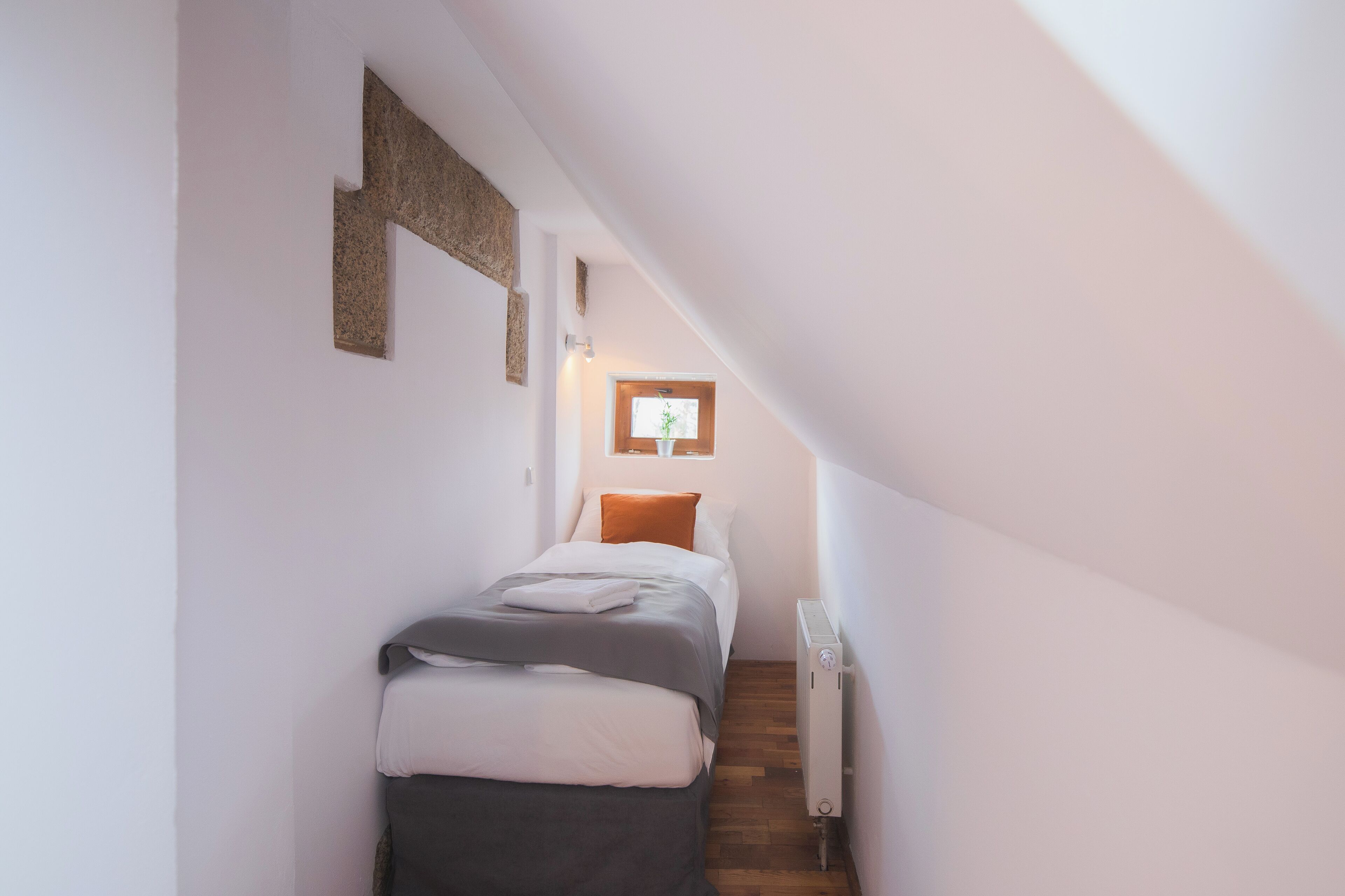 Een smalle, schuine kamer met een bed, een radiator en een raam met uitzicht.