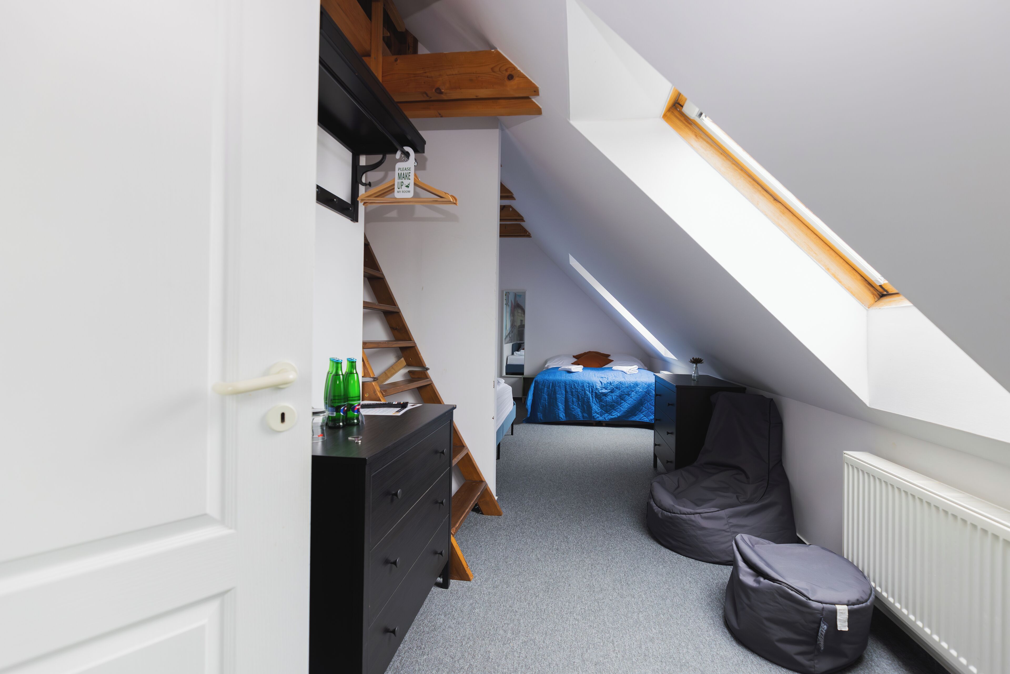 A narrow room with a sloped ceiling, a bed with a blue bedspread, a dresser, a chair, and a radiator.