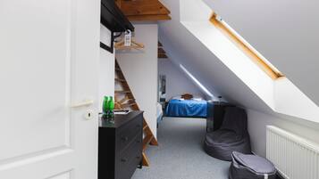 A narrow room with a sloped ceiling, a bed with a blue bedspread, a dresser, a chair, and a radiator.