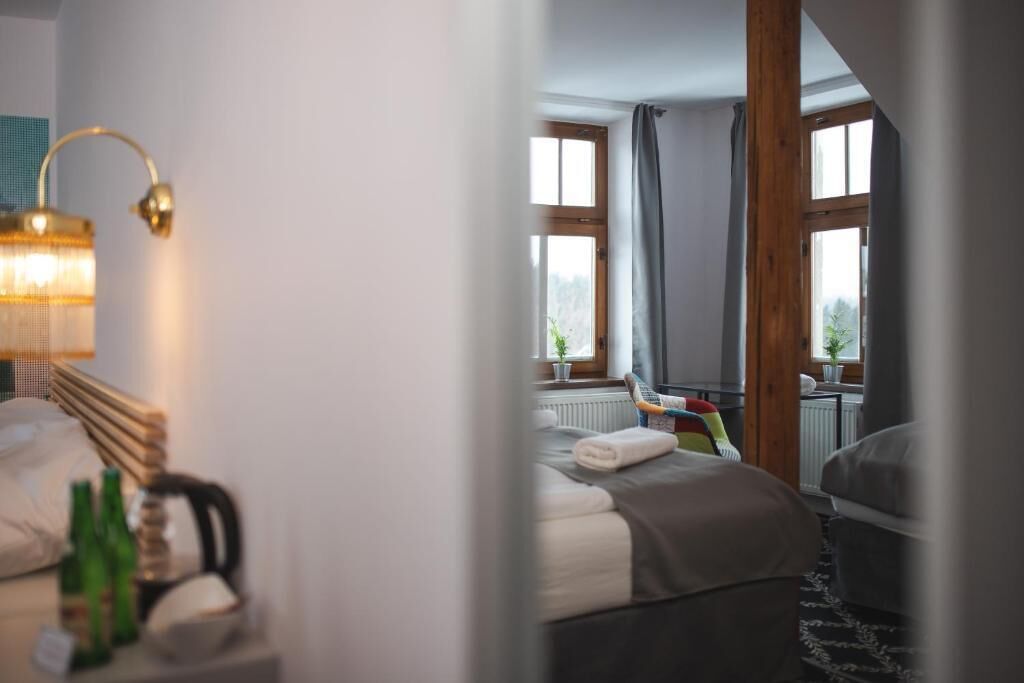 A hotel room with a bed, a nightstand with bottles, a lamp, and a view of the outdoors through large windows.