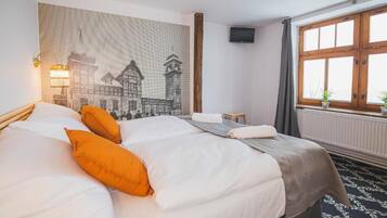 Une chambre d’hôtel avec un grand lit, deux oreillers, une tapisserie murale, une petite table, une chaise, une fenêtre avec des rideaux et un téléviseur.