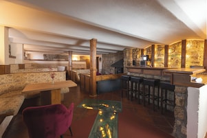Um espaço de bar acolhedor com parede de pedra, banquetas de madeira e área de estar com um sofá estampado e uma poltrona vermelha.