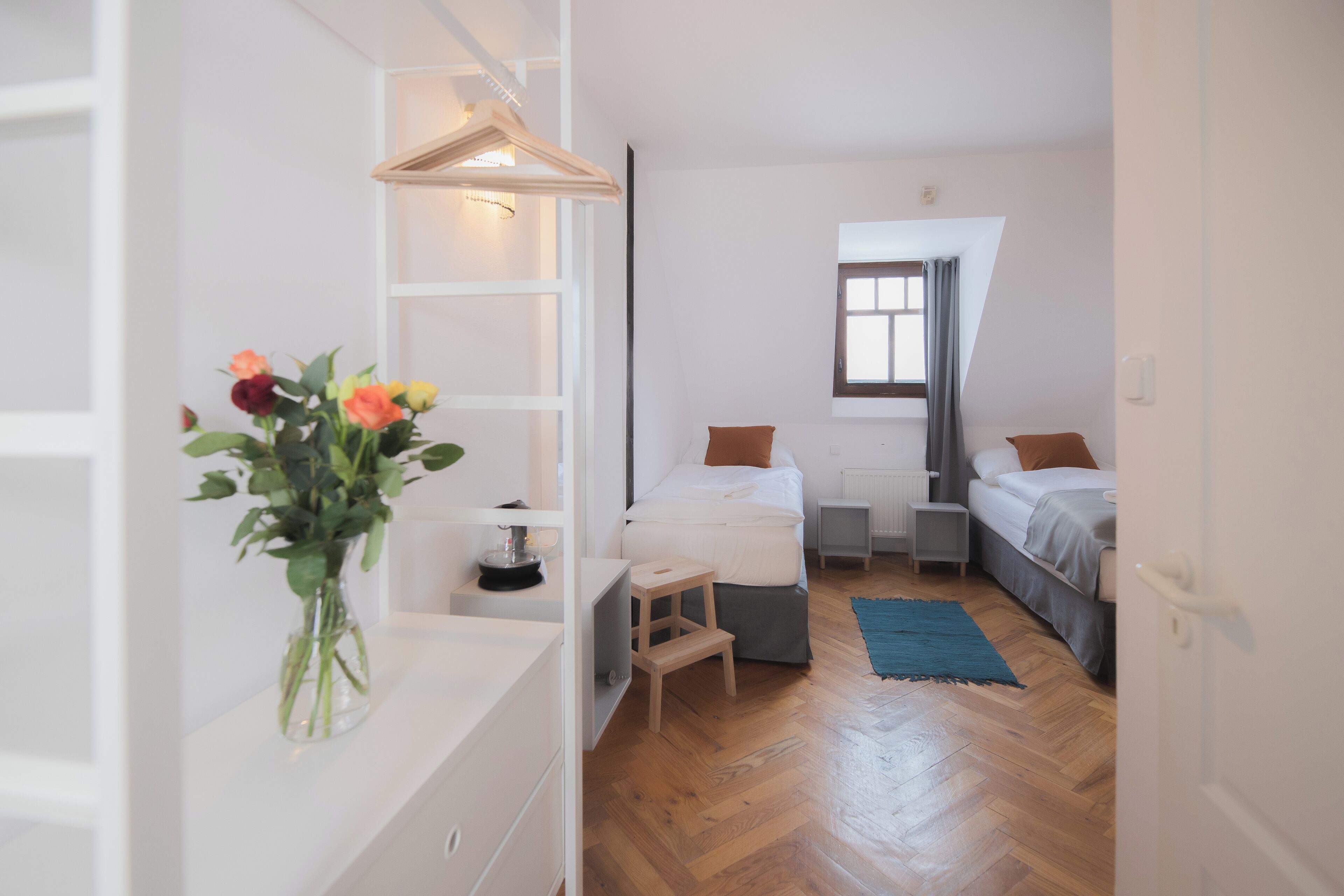 A modern bedroom with a bed, bedside tables, a small stool, a nightstand, a dresser, a hanging light fixture, and a window with curtains.