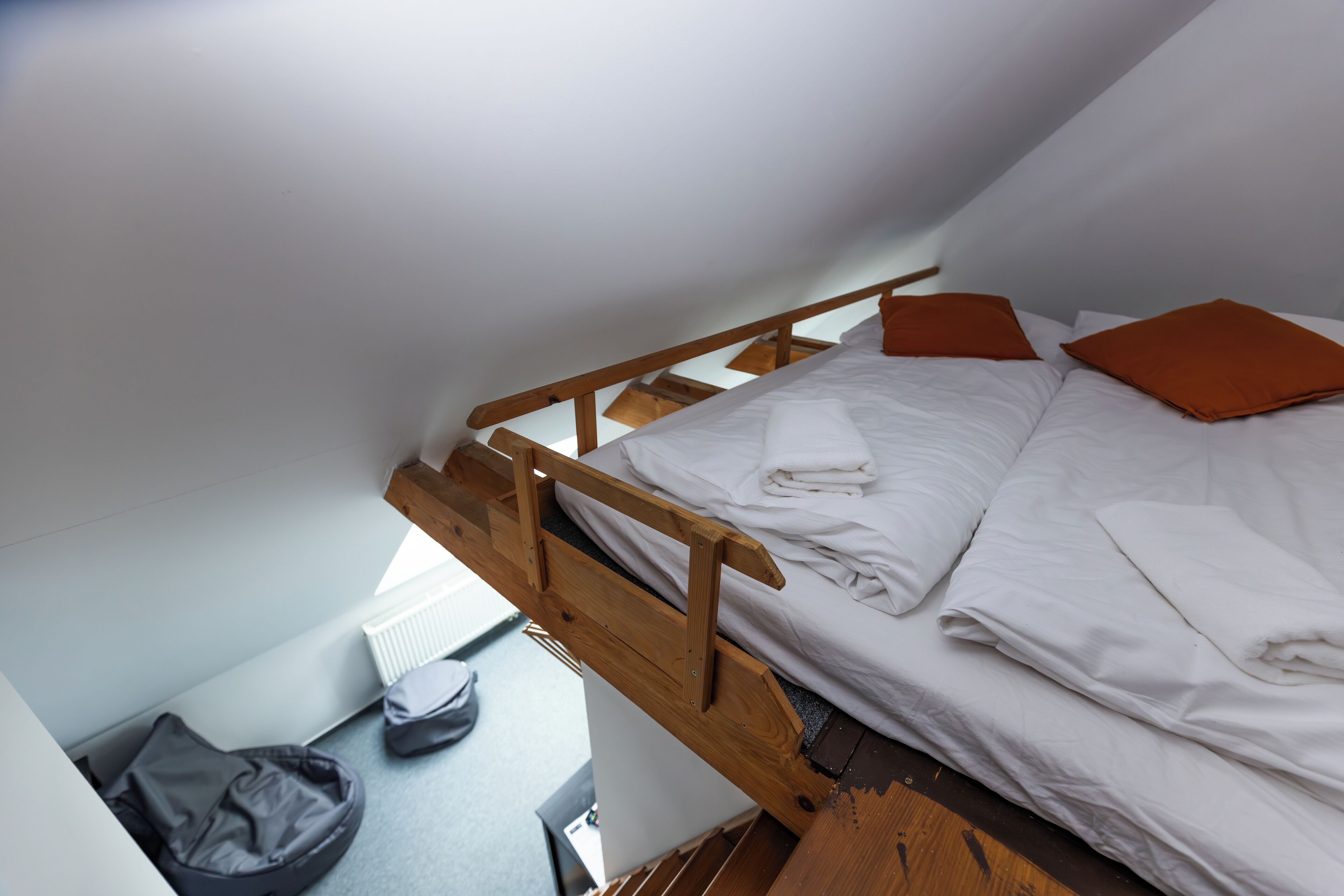 Un lit simple avec une tête de lit en bois et une literie blanche, comprenant un oreiller orange, dans une pièce dotée d’un plafond en pente et d’un radiateur.