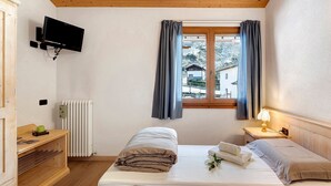 A room with two beds, a TV mounted on the wall, a window with curtains, a radiator, and a wooden shelf with items on it. - Room 'Camera Matrimoniale' with Mountain View, Shared Garden and Wi-Fi (Grosio)
