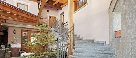 A stone staircase leading to a wooden upper level, a metal railing, and a small tree with snow on its branches.