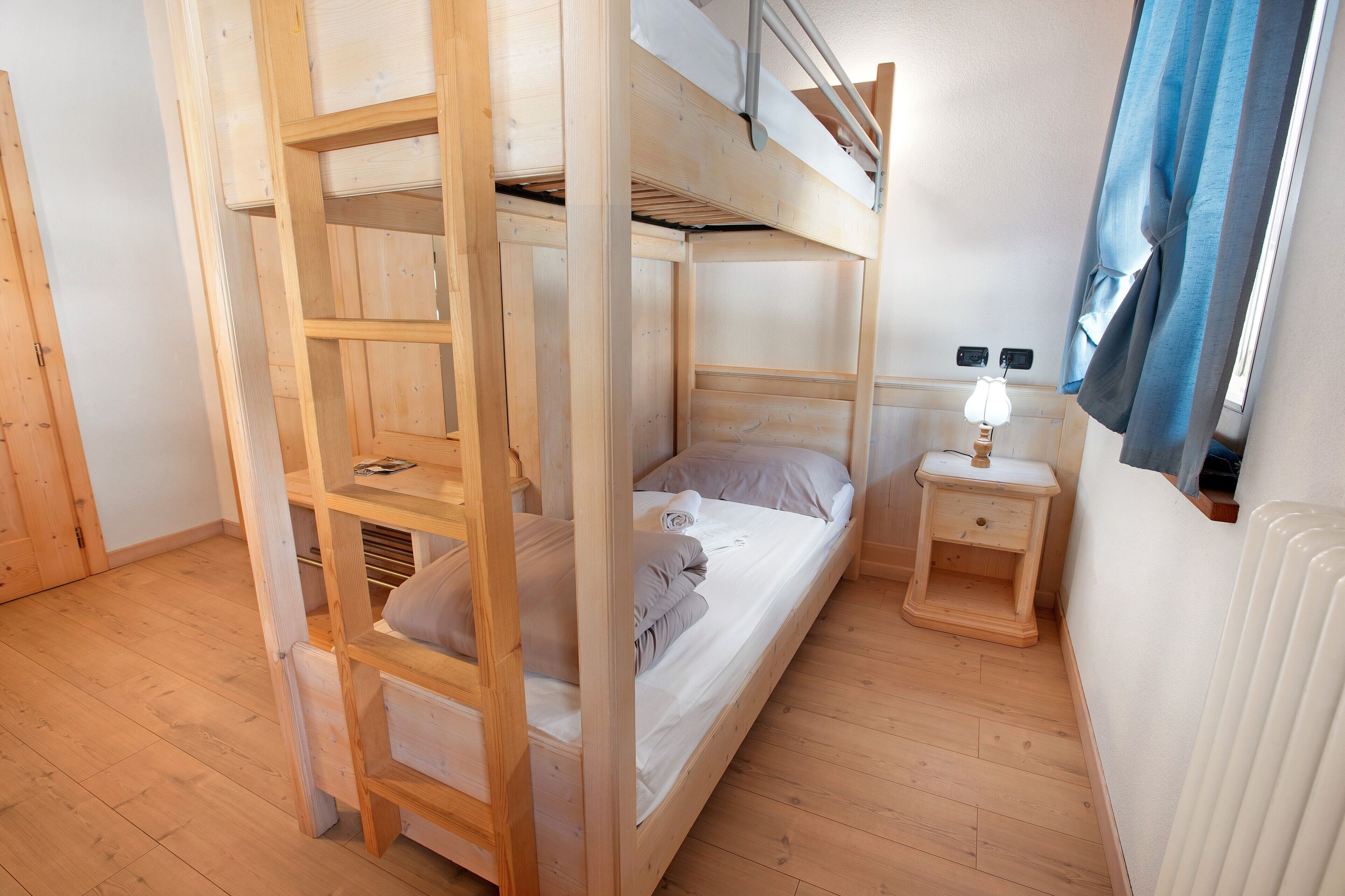 Een slaapkamer met een stapelbed, een houten ladder, een nachtkastje met lamp en een radiator.