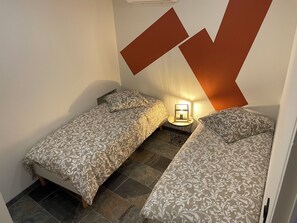 A bedroom with two single beds, a bedside table with a lamp, and a geometric wall design. - House in the Heart of the Village   (Cornillon)