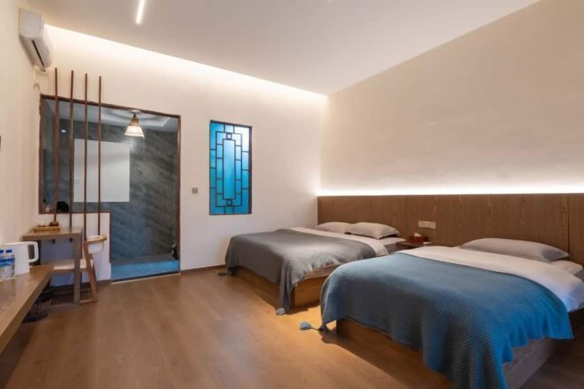 Quarto de hotel moderno com duas camas, uma escrivaninha de madeira, uma mesinha e uma obra de arte na parede.