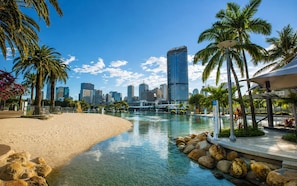 A beach with clear water, palm trees, and a city skyline in the background. - Relax & Unwind in Sunnybank Spacious 3BR Home w Parking (Sunnybank Hills)