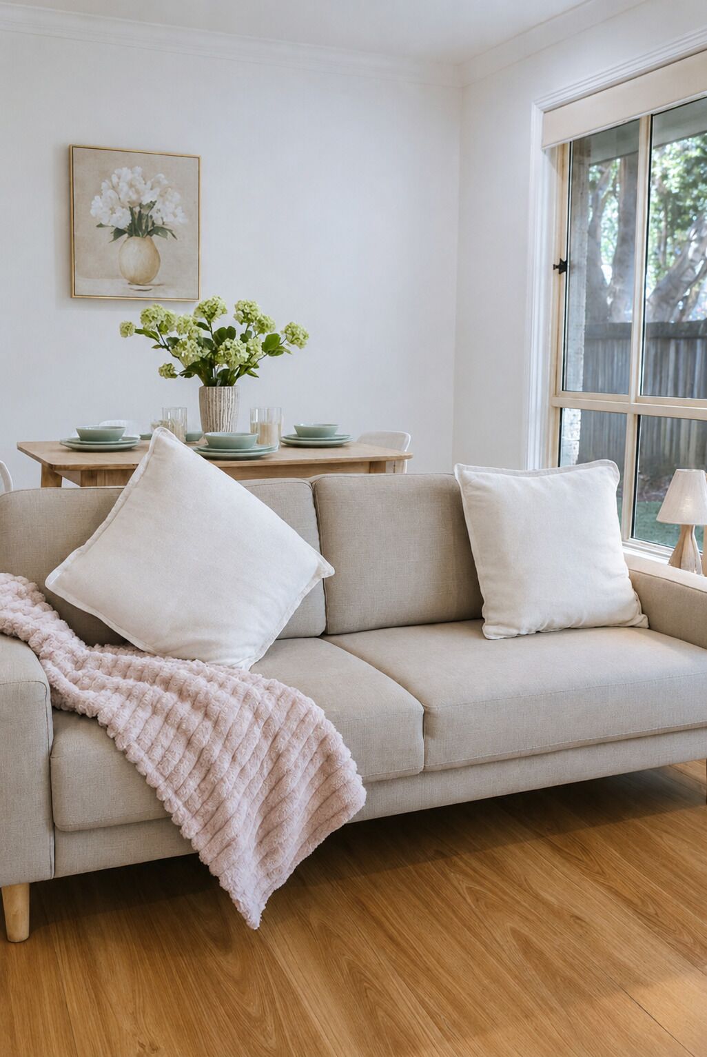 A living room with a beige sofa, a pink throw blanket, a dining table with plates and a vase of flowers, and a window with a view of trees.