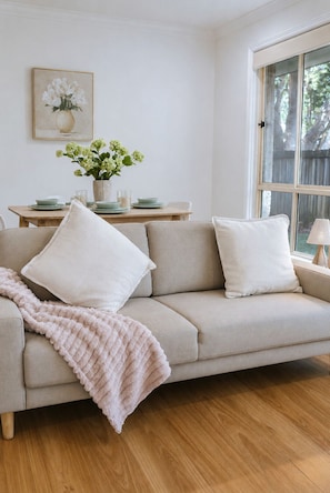 A living room with a beige sofa, a pink throw blanket, a dining table with plates and a vase of flowers, and a window with a view of trees.