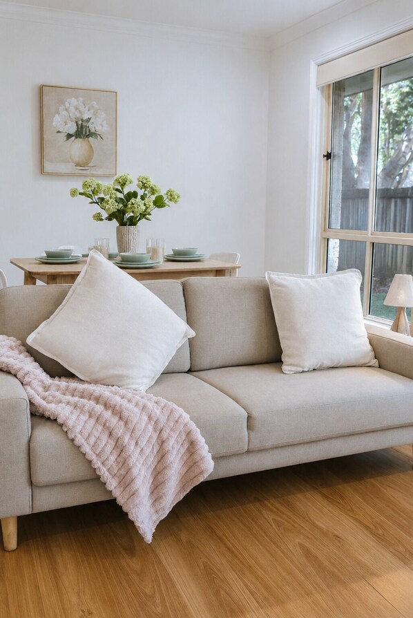 A living room with a beige sofa, a pink throw blanket, a dining table with plates and a vase of flowers, and a window with a view of trees.