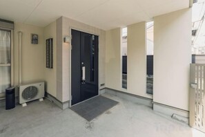 A hallway with a black door, a small air conditioning unit, and a mat at the entrance. - JS xiaozhai・yuishima (Osaka)