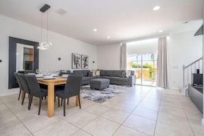 A modern living room with a dining area, grey sofas, a coffee table, and a large window with curtains. - Cozy, Perfect for Families, Hot Tub & Grill - 3013 (Kissimmee)