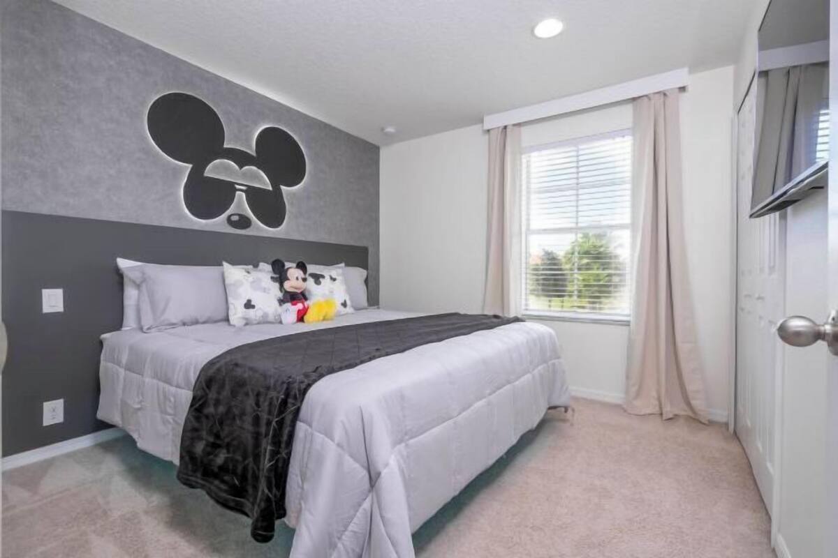 A bedroom with a large bed, a Mickey Mouse wall decoration, a window with curtains, and a door with a handle.