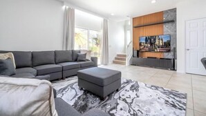 A modern living room with a sectional sofa, ottoman, and a large flat-screen TV mounted on a wooden wall.