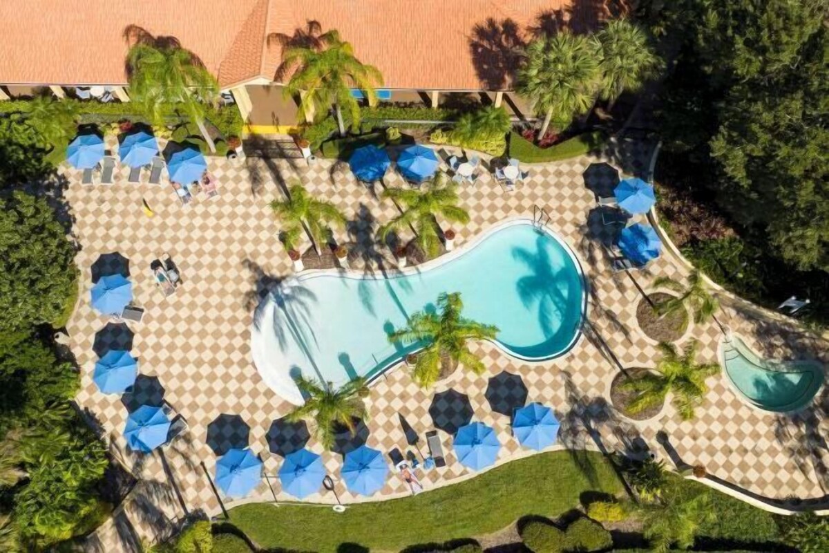 An aerial view of a resort with a central swimming pool, surrounded by checkered lounge areas with umbrellas and palm trees.