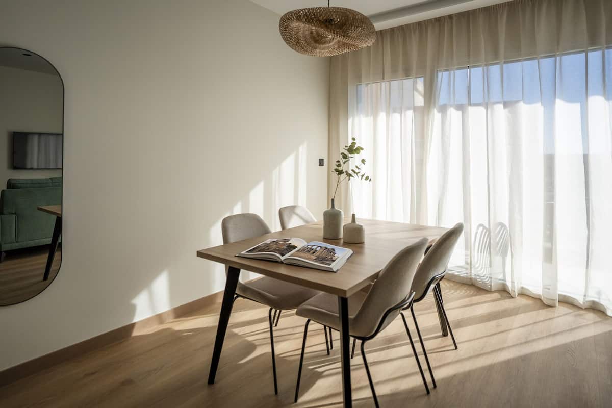 A dining area with a wooden table, four chairs, a hanging light fixture, and a large window with sheer curtains.
