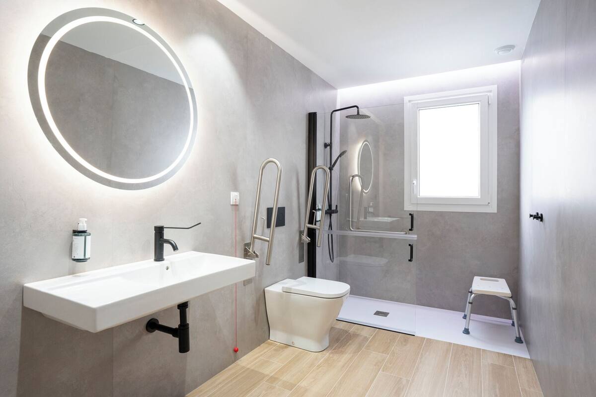 A modern bathroom with a white sink, a round mirror, a toilet, a shower, and a small step stool.