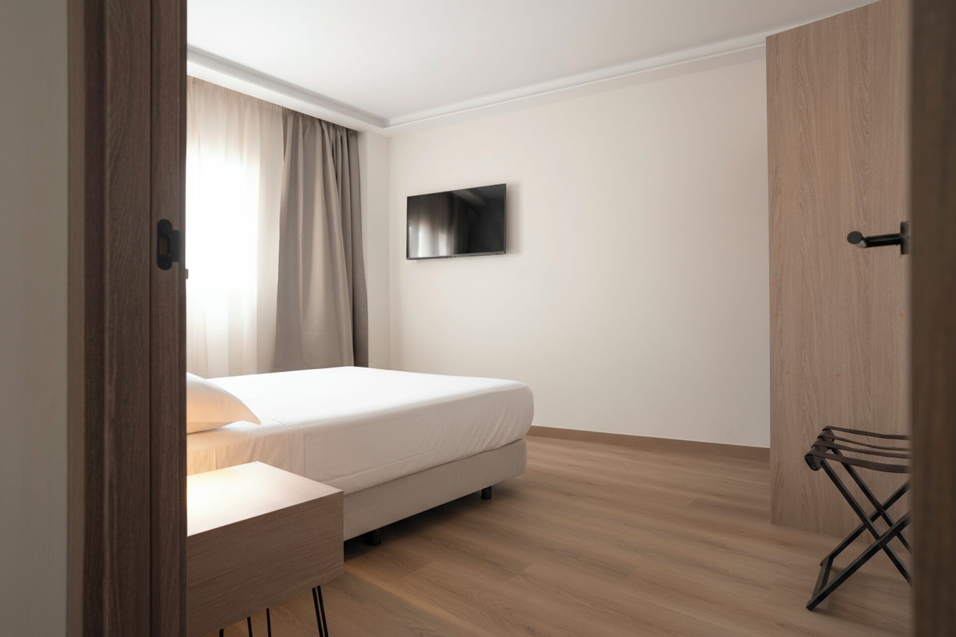 Una habitación de hotel minimalista con una cama, un televisor montado en la pared y suelo de madera.